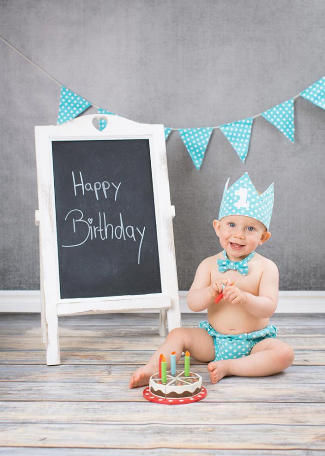 Junge sitzt am ersten Geburtstag in Partyoutfit vor Holzkuchen und Tafel