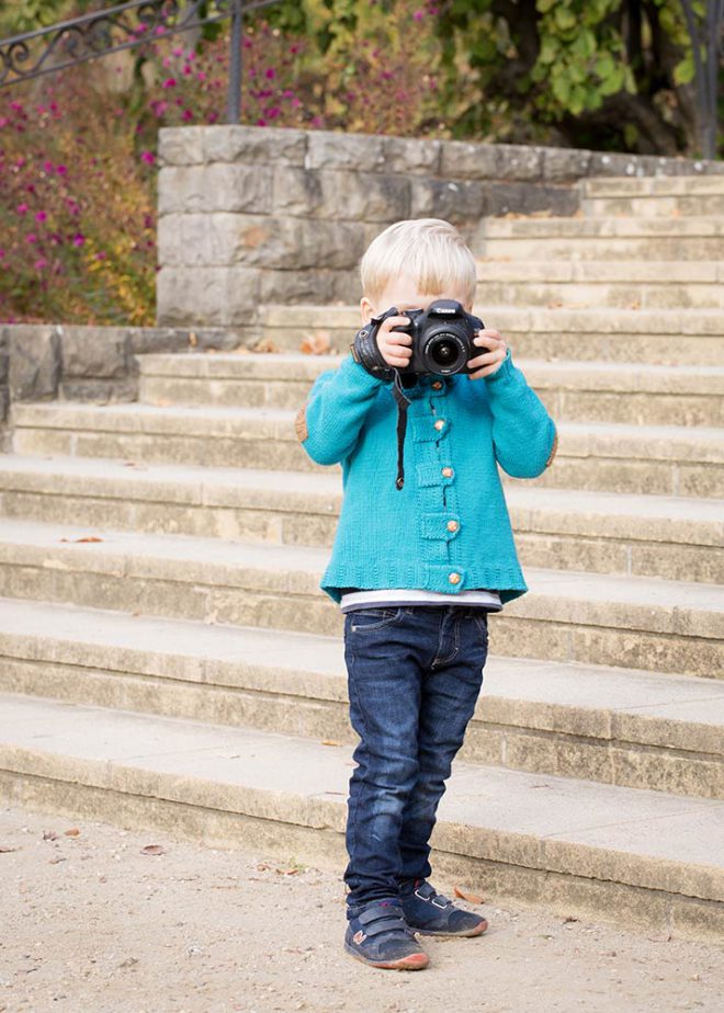 Junge mit blauer Jacke steht mit Kamera vor einer Treppe